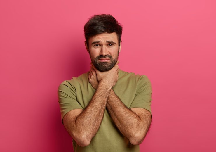 Unshaven displeased man keeps hands on neck, suffers from suffocation, has sore throat, irritation, being unhealthy, looks unhappily at camera, stands against pink background, hard to swallow